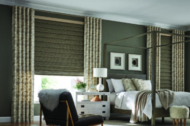 Natural, wooden woven shades with patterned drapes framing each side in a home bedroom. Available at The Blind Broker of Kansas City.