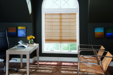 A set of wooden woven shades adorning a large, arch-shaped window in a home office. Available at The Blind Broker of Kansas City.