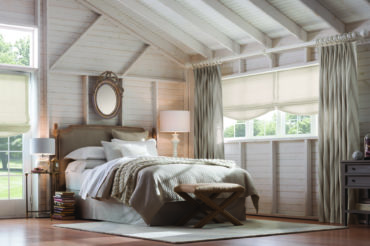 Custom natural-tone drapes in a brightly-lit bedroom, framing roman shades in a natural tone. Available at The Blind Broker of Kansas City.
