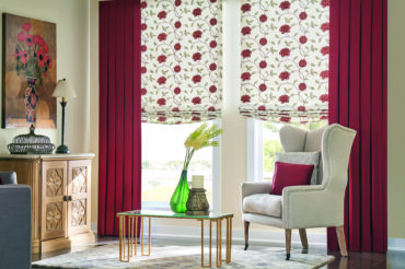 Artisan red drapes accenting floral pattern roman shades in a contemporary living space with ample natural light. Styles available at The Blind Broker of Kansas City.